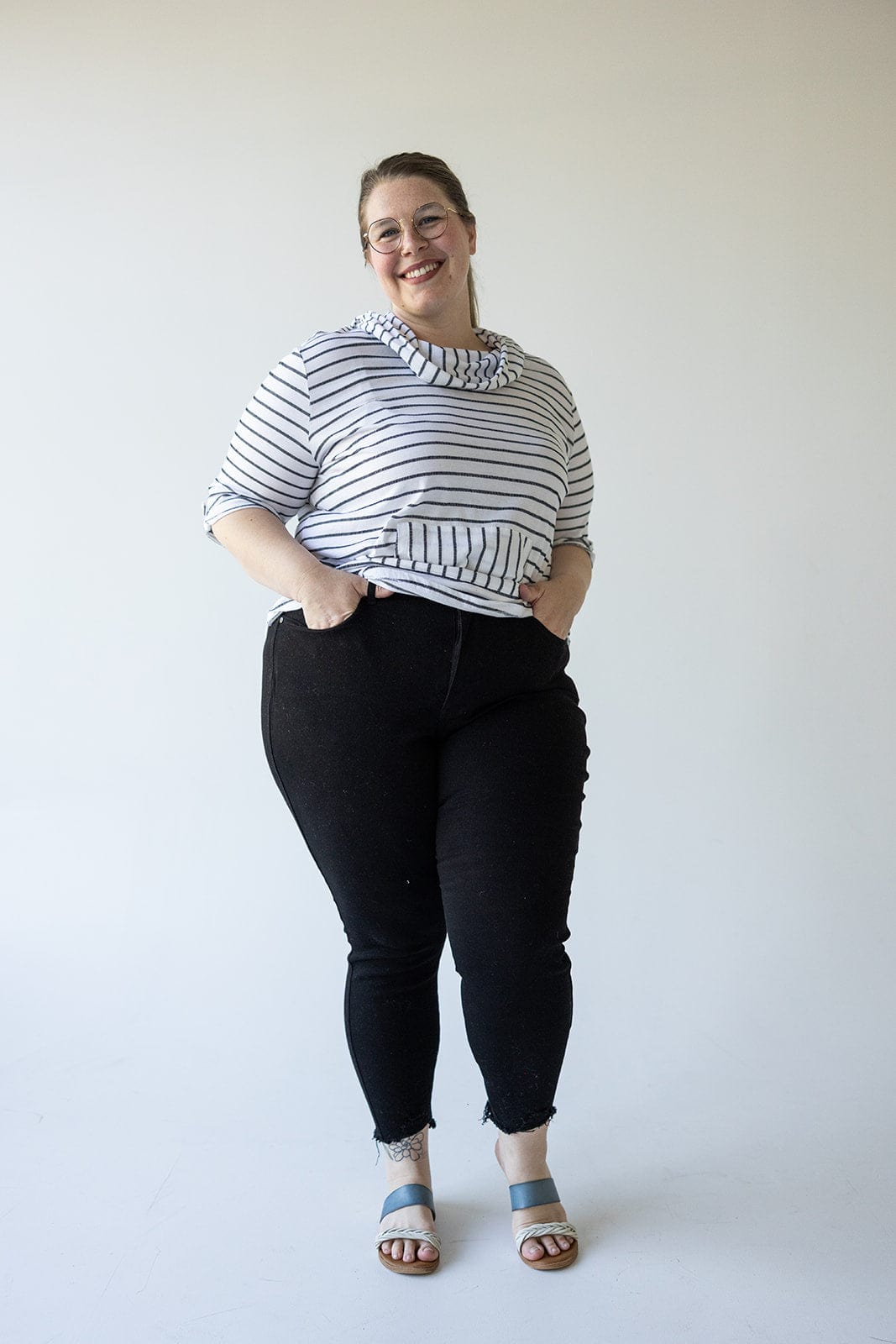 A person stands against a plain background, wearing a striped top, Judy Blue HIGH WAIST TUMMY CONTROL SKINNY JEANS WITH DISTRESSED HEM IN BLACK, and sandals, smiling with hands in pockets.