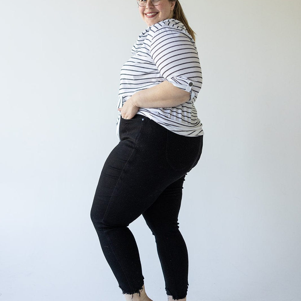 A woman wearing a striped shirt, Judy Blue High Waist Tummy Control Skinny Jeans with Distressed Hem in Black, and blue sandals stands facing left, smiling.