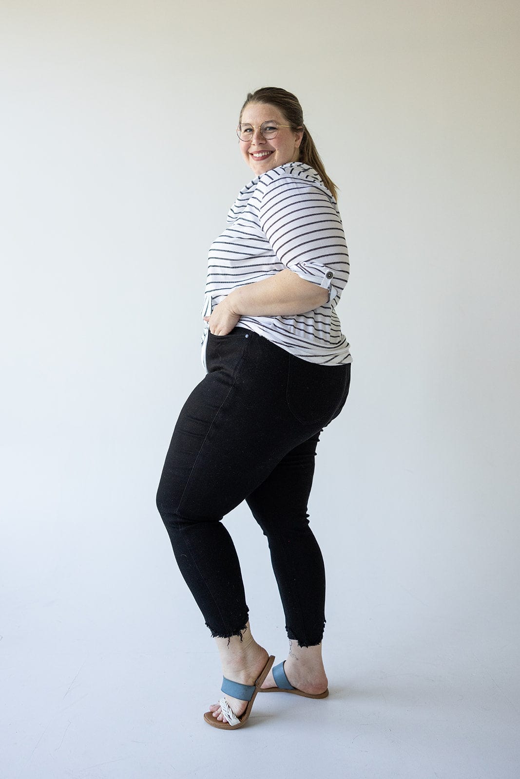 A woman wearing a striped shirt, Judy Blue High Waist Tummy Control Skinny Jeans with Distressed Hem in Black, and blue sandals stands facing left, smiling.