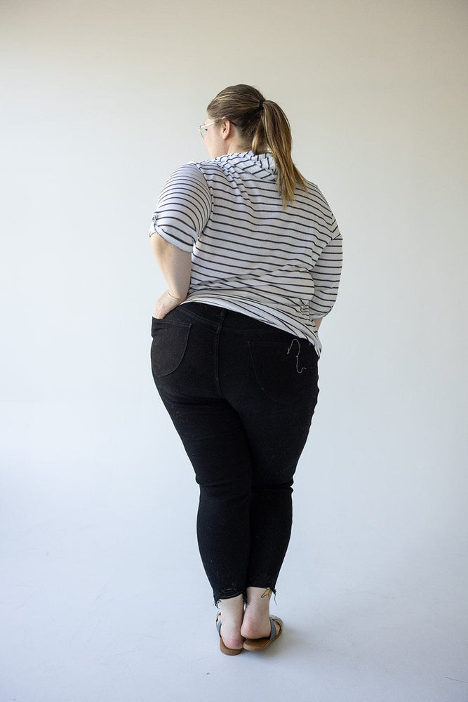 A person stands facing away from the camera, wearing a striped shirt, sandals, and Judy Blue High Waist Tummy Control Skinny Jeans with Distressed Hem in Black by Judy Blue, with their hair tied in a ponytail. The background is plain and light-colored.