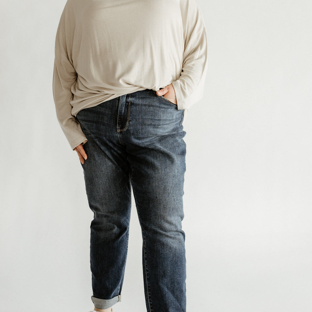 A person stands confidently in beige long-sleeve top, Judy Blue jeans, and white sneakers, hands in pockets against a plain background.