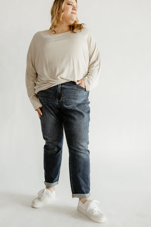 A person stands confidently in beige long-sleeve top, Judy Blue jeans, and white sneakers, hands in pockets against a plain background.