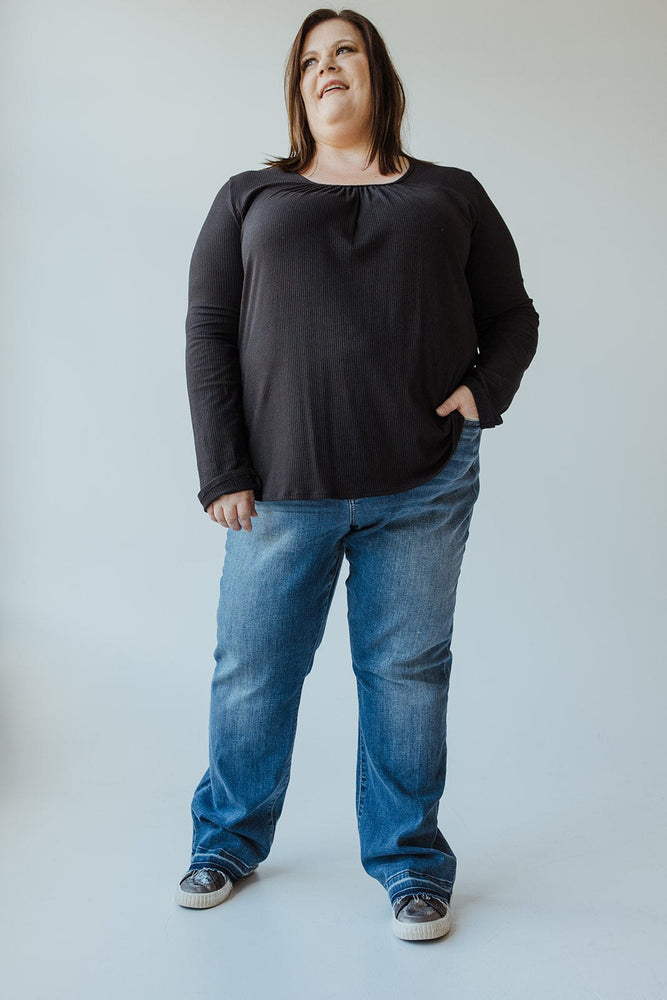 A person stands against a plain background, wearing a long-sleeve black shirt, Judy Blue Mid-Rise Dad Jeans with Released Hem, and sneakers. They are smiling and have one hand in their pocket.