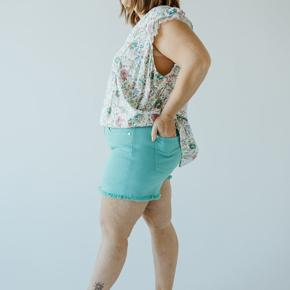 A person with shoulder-length hair wearing a floral sleeveless top, Judy Blue JUDY BLUE MID-RISE FRAYED HEM SHORTS IN MERMAID, and beige sandals stands in profile against a plain background.