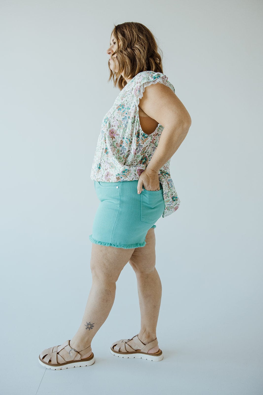 A person with shoulder-length hair wearing a floral sleeveless top, Judy Blue JUDY BLUE MID-RISE FRAYED HEM SHORTS IN MERMAID, and beige sandals stands in profile against a plain background.