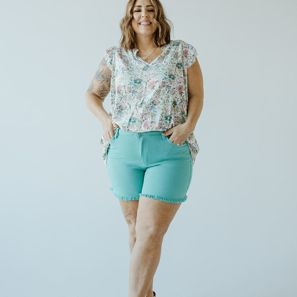 A person stands smiling against a plain background, dressed in a floral top and Judy Blue JUDY BLUE MID-RISE FRAYED HEM SHORTS IN MERMAID that accentuate their style. They complete the look with sandals, hands casually tucked in their pockets.