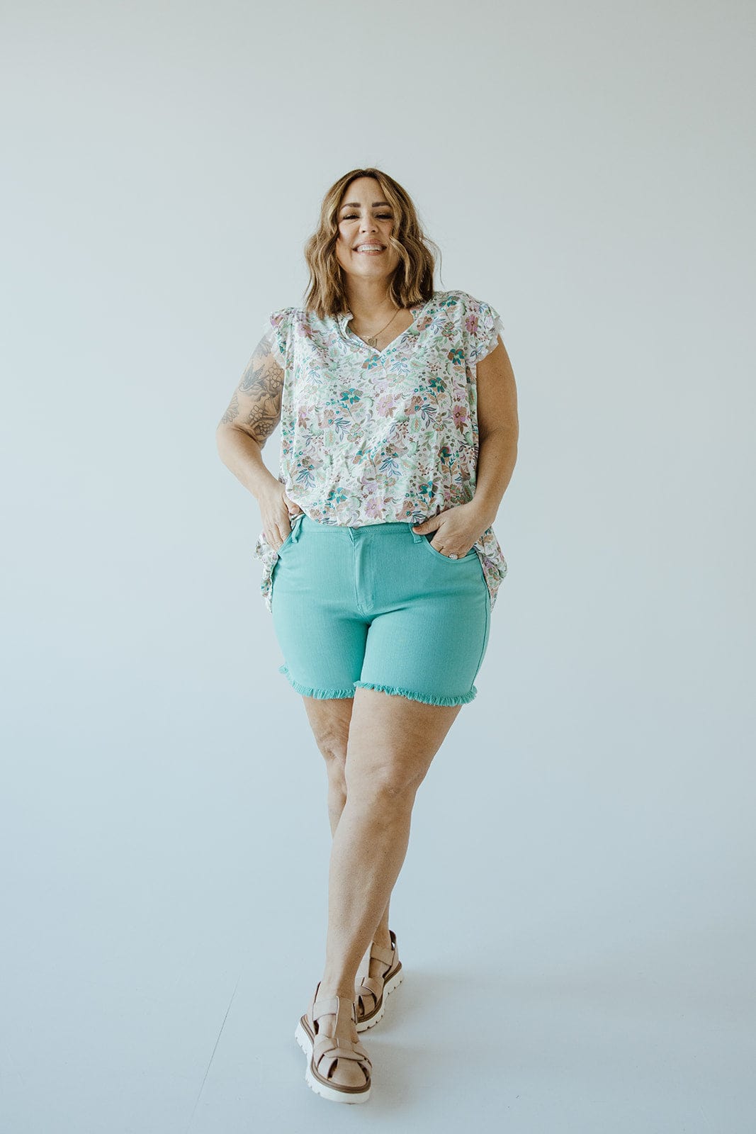 A person stands smiling against a plain background, dressed in a floral top and Judy Blue JUDY BLUE MID-RISE FRAYED HEM SHORTS IN MERMAID that accentuate their style. They complete the look with sandals, hands casually tucked in their pockets.