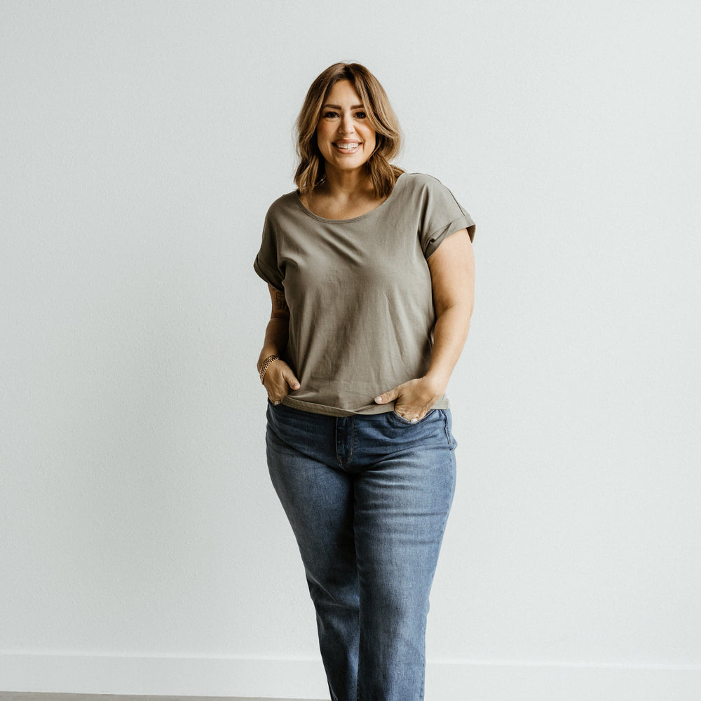 A person stands against a plain background, smiling, wearing a gray t-shirt, Judy Blue MID-RISE BOYFRIEND JEANS in dark wash, and black shoes.