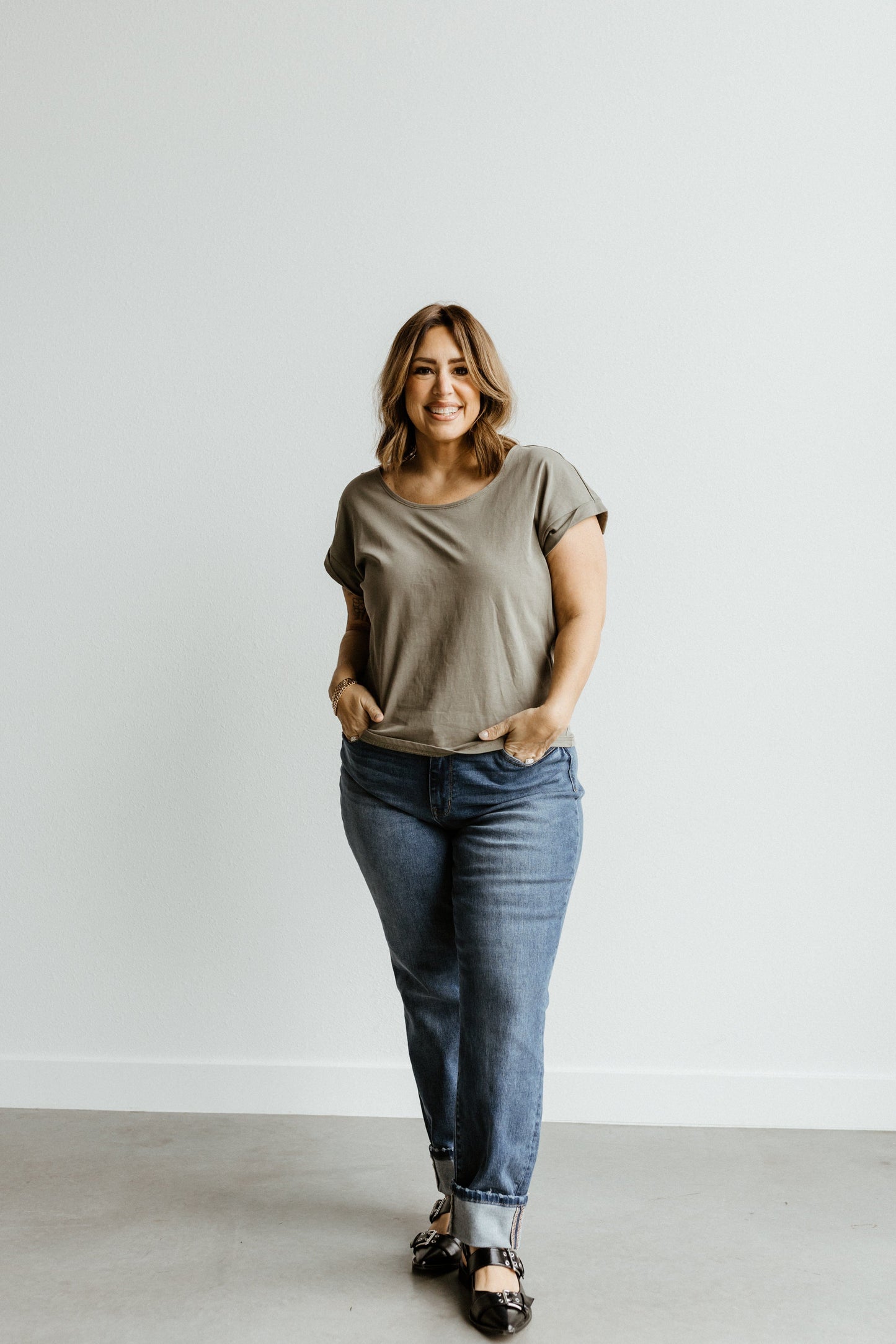 A person stands against a plain background, smiling, wearing a gray t-shirt, Judy Blue MID-RISE BOYFRIEND JEANS in dark wash, and black shoes.