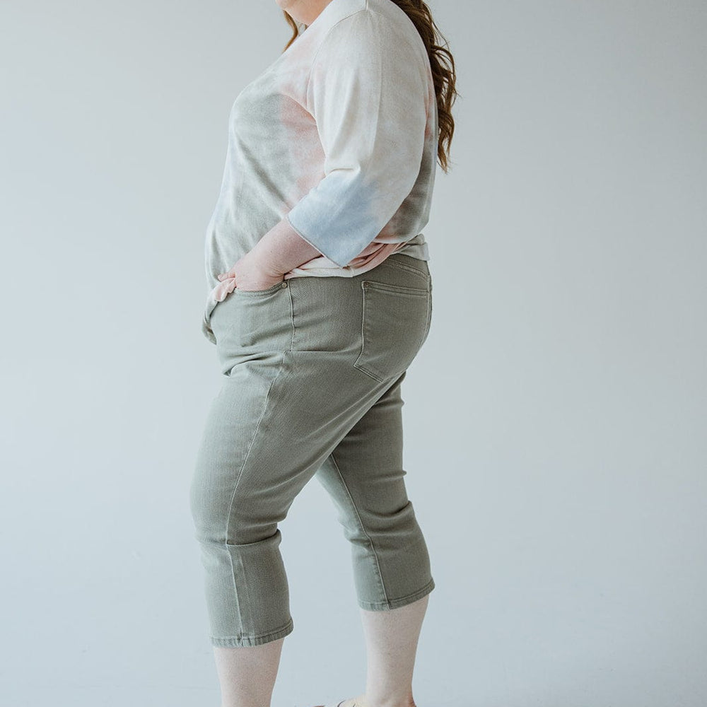 A person with long hair, wearing a light-colored top, Judy Blue Mid-Rise Non-Distressed Capri in Sage from Judy Blue, and beige sandals, stands sideways with hands in pockets.