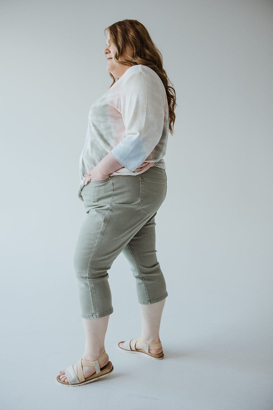 A person with long hair, wearing a light-colored top, Judy Blue Mid-Rise Non-Distressed Capri in Sage from Judy Blue, and beige sandals, stands sideways with hands in pockets.