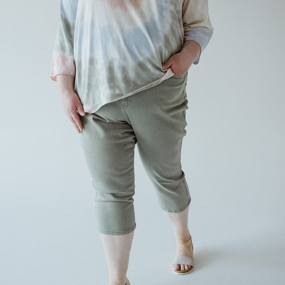 A person is wearing a pastel tie-dye shirt, Judy Blue Mid-Rise Non-Distressed Capris in Sage by Judy Blue, and beige sandals. The person's left hand is in their pocket.