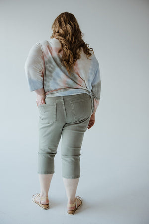 A person with long hair stands facing away, wearing a tie-dye shirt, Judy Blue Mid-Rise Non-Distressed Capri in Sage, and sandals.