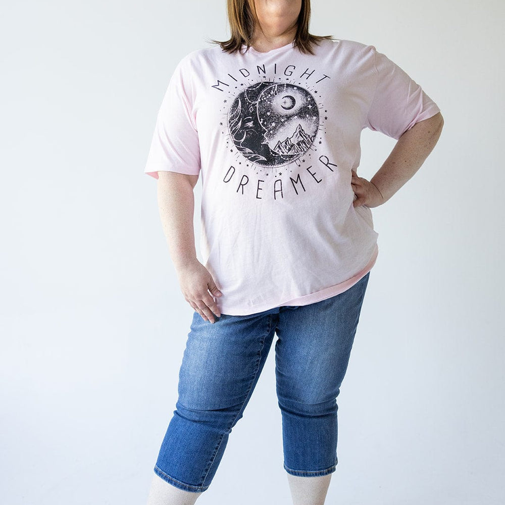 A woman stands against a plain background, wearing a pink t-shirt with "Midnight Dreamer" text and graphic, Judy Blue MID-RISE NON-DISTRESSED CAPRI WITH SIDE SLIT jeans, and black sandals. She has her hand on her hip and is looking off-camera.