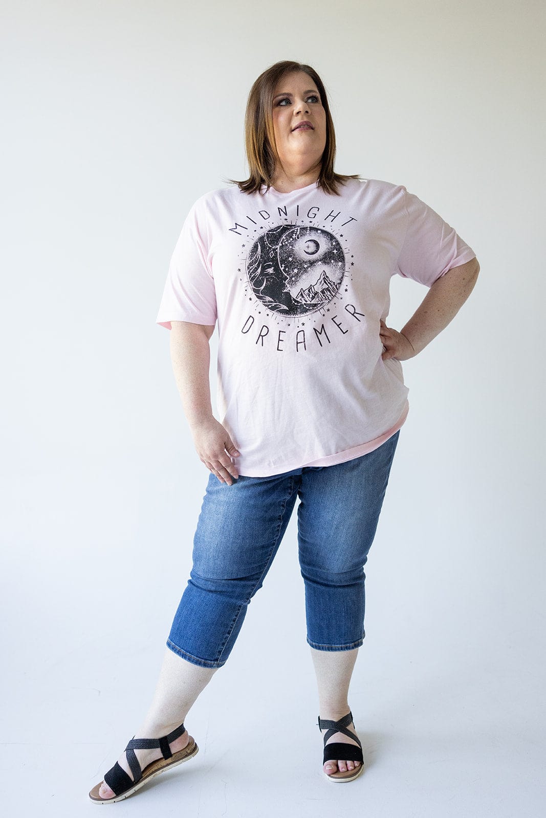 A woman stands against a plain background, wearing a pink t-shirt with "Midnight Dreamer" text and graphic, Judy Blue MID-RISE NON-DISTRESSED CAPRI WITH SIDE SLIT jeans, and black sandals. She has her hand on her hip and is looking off-camera.