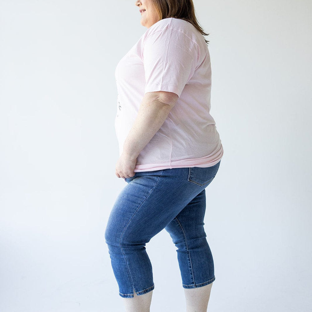 A person is standing in profile, smiling, wearing a pink t-shirt, Judy Blue Mid-Rise Non-Distressed Capri with Side Slit jeans, and black sandals against a plain white background.