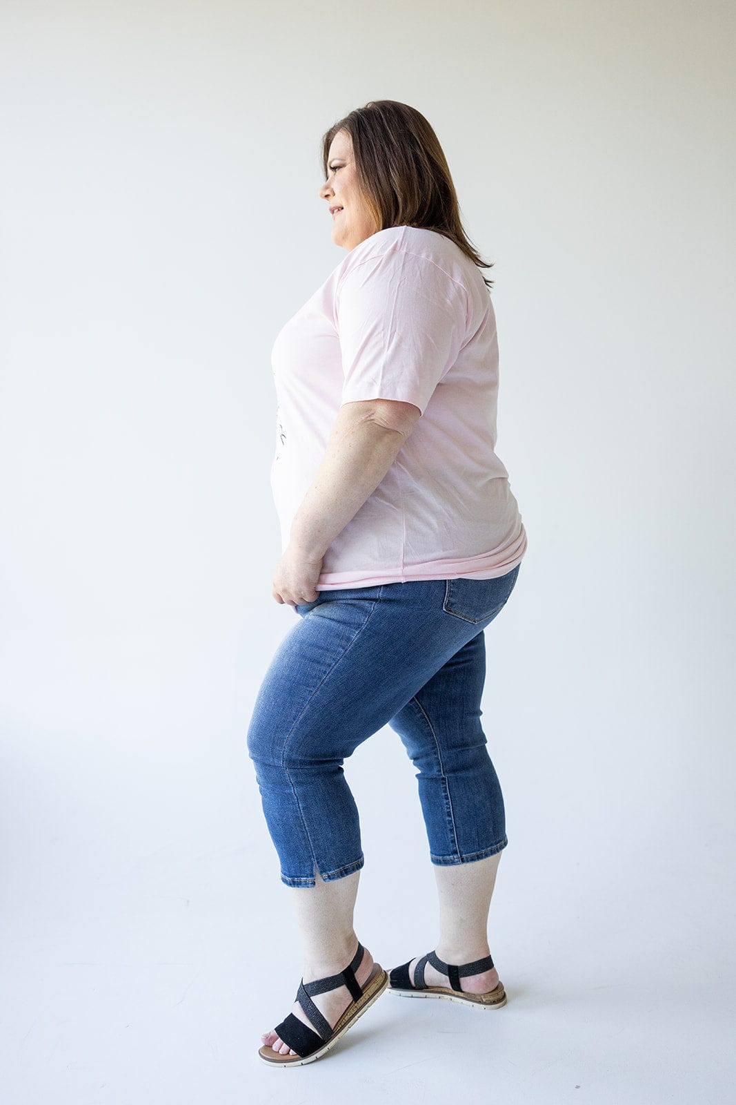 A person is standing in profile, smiling, wearing a pink t-shirt, Judy Blue Mid-Rise Non-Distressed Capri with Side Slit jeans, and black sandals against a plain white background.