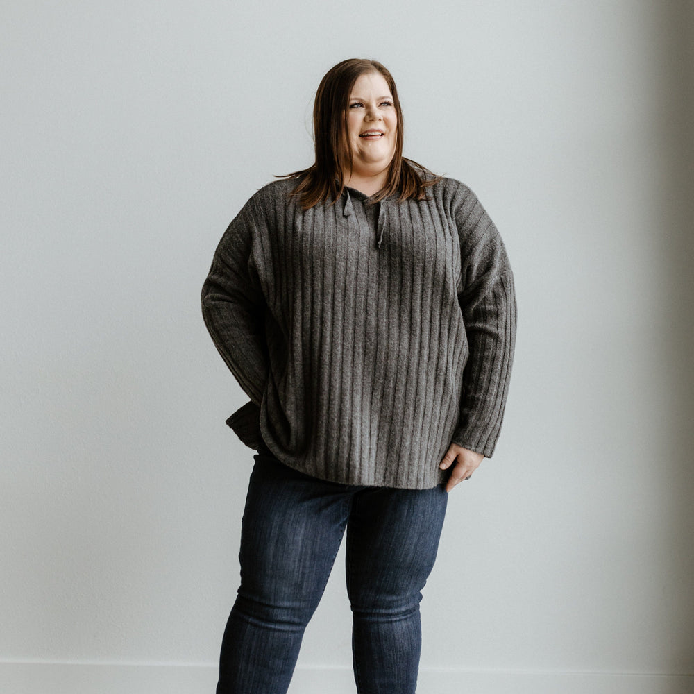 A person stands smiling against a plain, light-colored wall, wearing a gray sweater, Judy Blue mid-rise skinny jeans in a dark wash, and white sneakers.