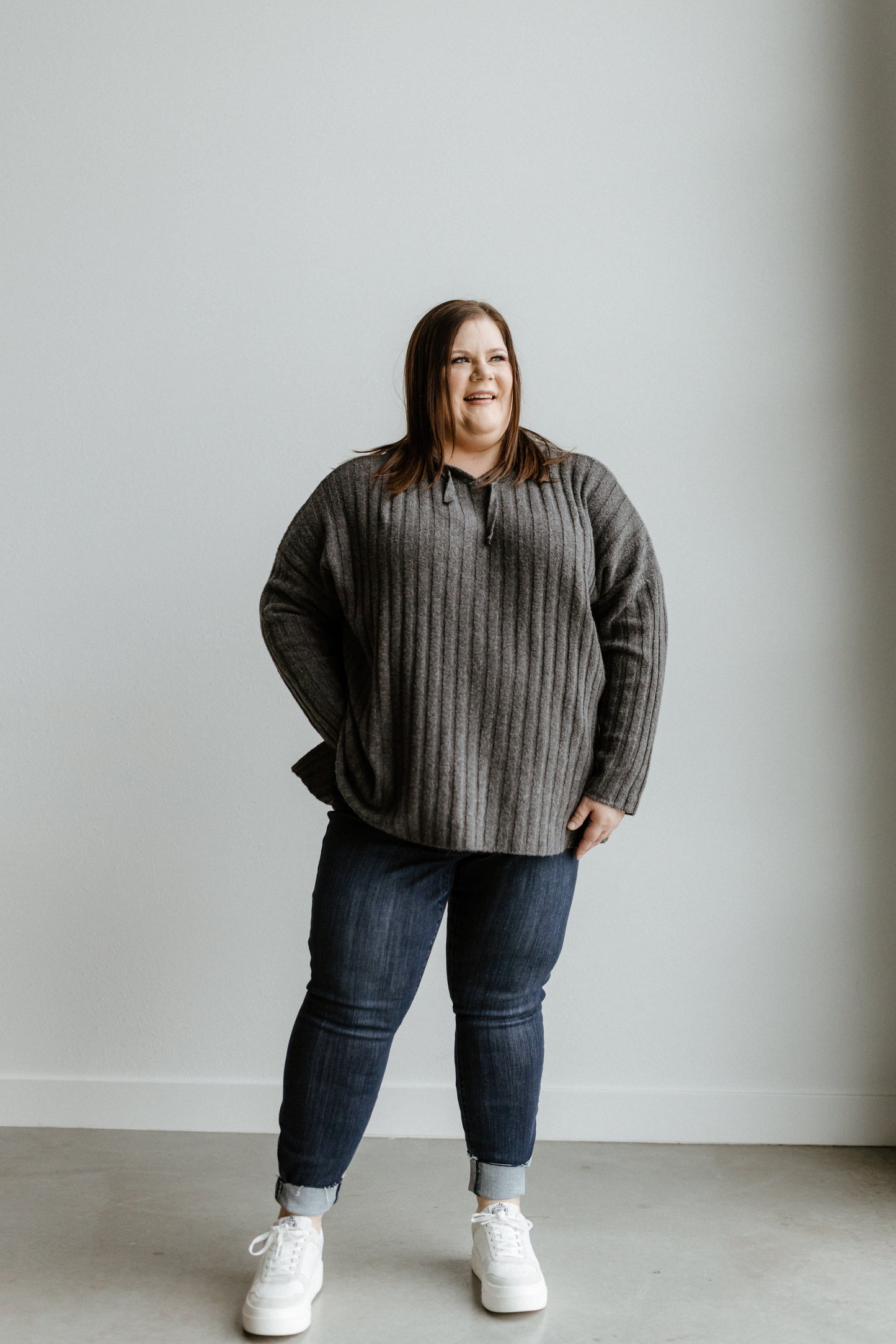 A person stands smiling against a plain, light-colored wall, wearing a gray sweater, Judy Blue mid-rise skinny jeans in a dark wash, and white sneakers.