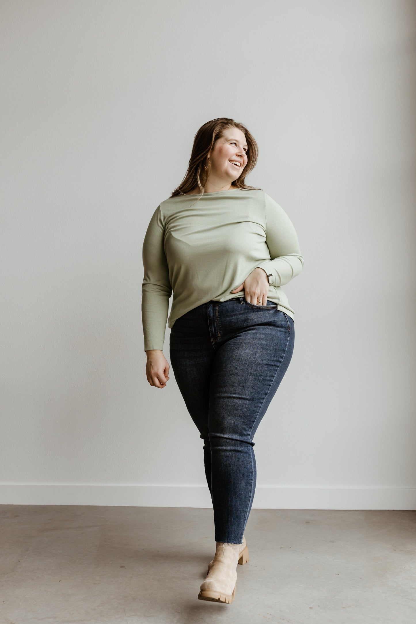 A person stands against a plain wall, wearing a light green long-sleeve top and mid-rise skinny jeans paired with beige heels, exuding a relaxed posture and a slight smile.