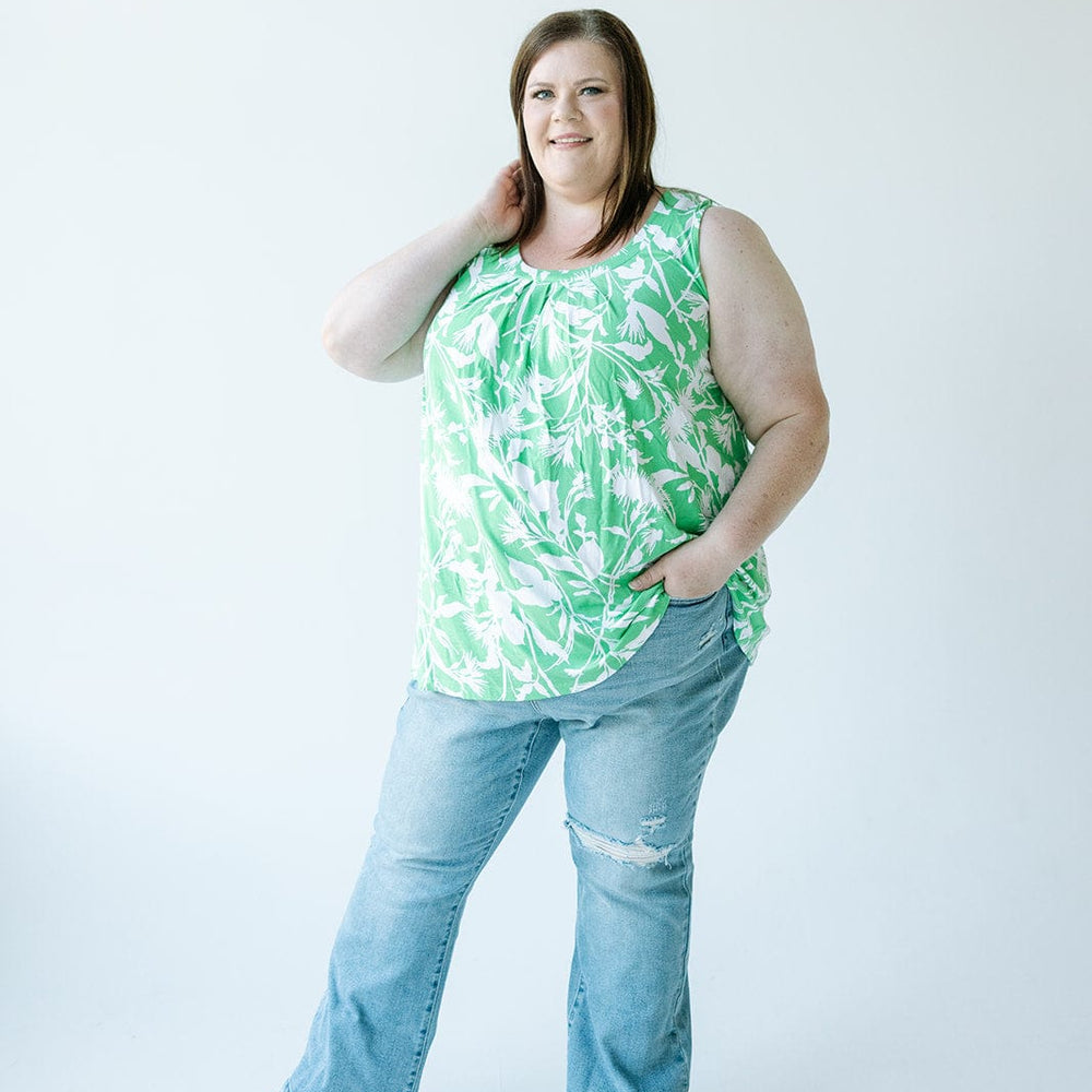 A person in a green floral top and Judy Blue bootcut jeans stands against a plain background, with one hand on their neck and the other in a pocket.