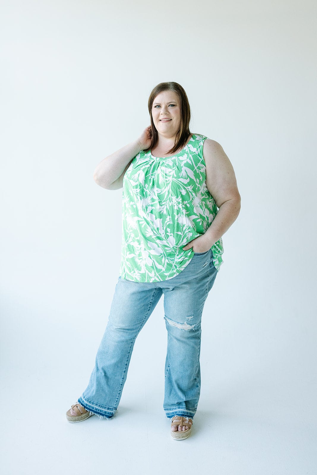 A person in a green floral top and Judy Blue bootcut jeans stands against a plain background, with one hand on their neck and the other in a pocket.