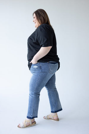 A person is standing sideways with hands in pockets, wearing a black T-shirt, JUDY BLUE MID-RISE RELEASED HEM CROPPED BOOTCUT jeans by Judy Blue, and beige sandals against a plain white background.