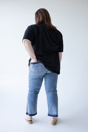 A person with brown hair wearing a black shirt, Judy Blue JUDY BLUE MID-RISE RELEASED HEM CROPPED BOOTCUT jeans, and sandals is standing with their back to the camera, one hand in their pocket.