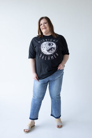 A person is standing against a plain white background, smiling, wearing a black "Midnight Dreamer" shirt, JUDY BLUE MID-RISE RELEASED HEM CROPPED BOOTCUT jeans by Judy Blue, and tan sandals.