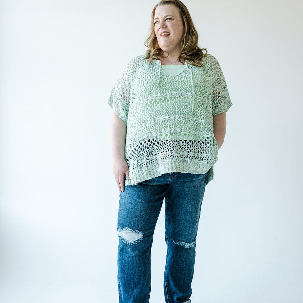 A woman standing against a plain white background, wearing a light green crocheted top over a green undershirt, Judy Blue Mid-Rise Slim Straight Rolled Cuff Jeans with holes, and black and beige sandals.