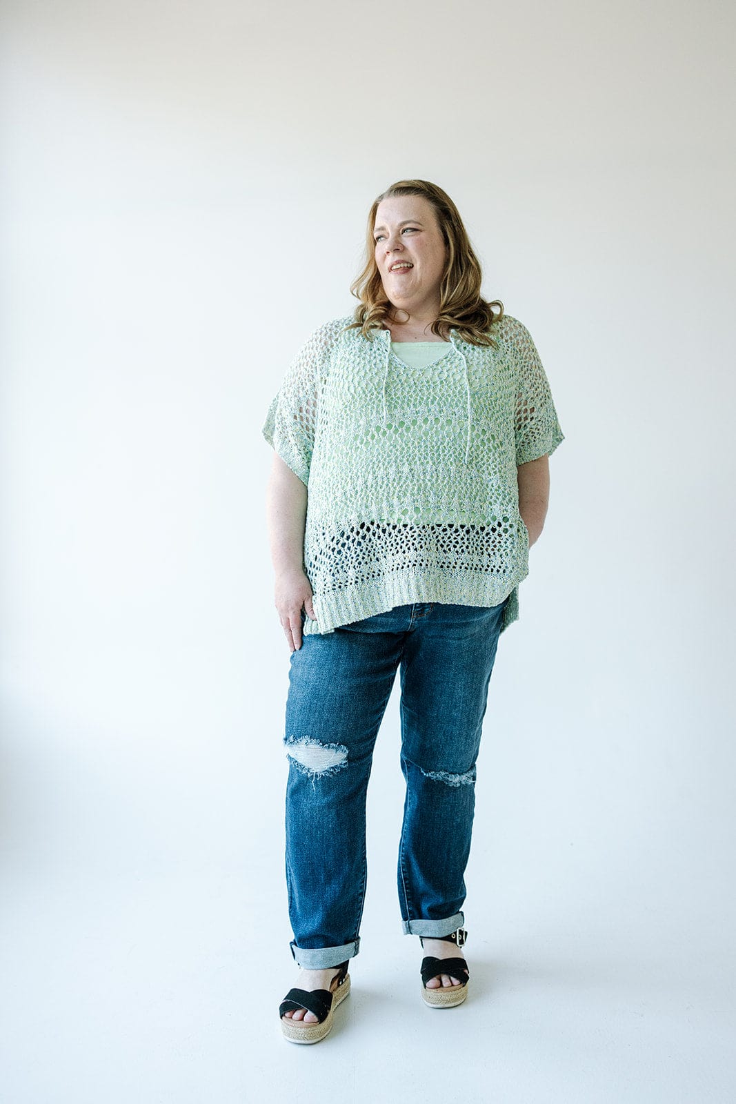 A woman standing against a plain white background, wearing a light green crocheted top over a green undershirt, Judy Blue Mid-Rise Slim Straight Rolled Cuff Jeans with holes, and black and beige sandals.