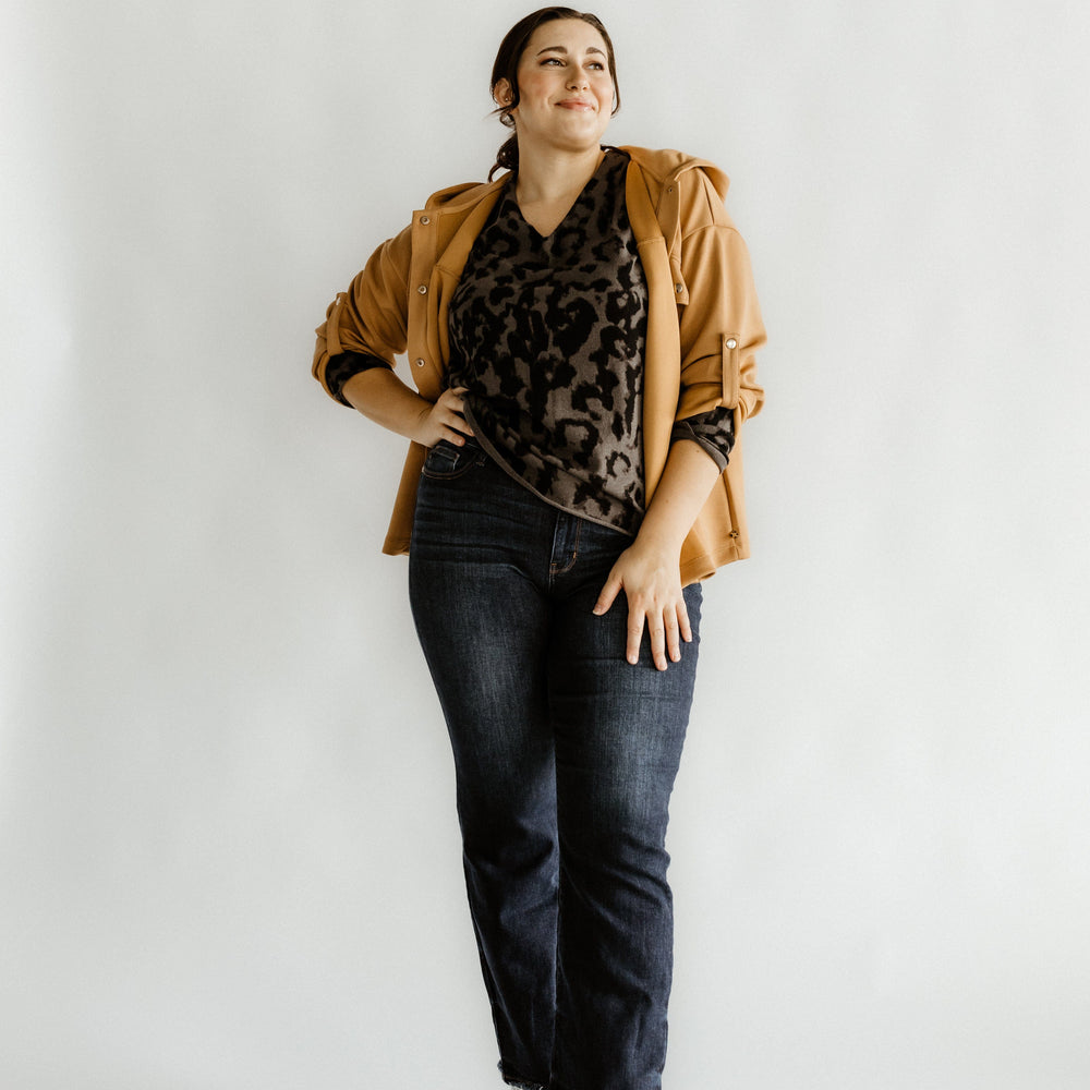 Person posing confidently in a leopard print top, brown jacket, and Judy Blue mid-rise straight leg jeans paired with black boots against a plain background.