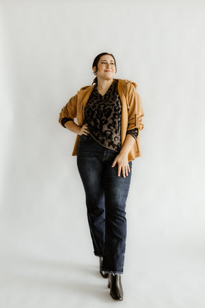 Person posing confidently in a leopard print top, brown jacket, and Judy Blue mid-rise straight leg jeans paired with black boots against a plain background.