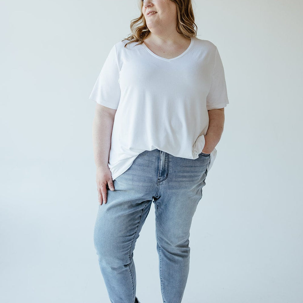 A woman is standing against a plain white background, wearing a white t-shirt, Judy Blue JUDY BLUE MID-RISE SUSTAINABLE DENIM IN A RELAXED FIT WITH COOLING TECHNOLOGY, and brown shoes, with her hands in her pockets. She is looking off to the side.