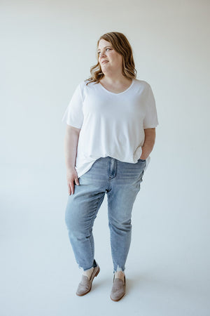 A woman is standing against a plain white background, wearing a white t-shirt, Judy Blue JUDY BLUE MID-RISE SUSTAINABLE DENIM IN A RELAXED FIT WITH COOLING TECHNOLOGY, and brown shoes, with her hands in her pockets. She is looking off to the side.