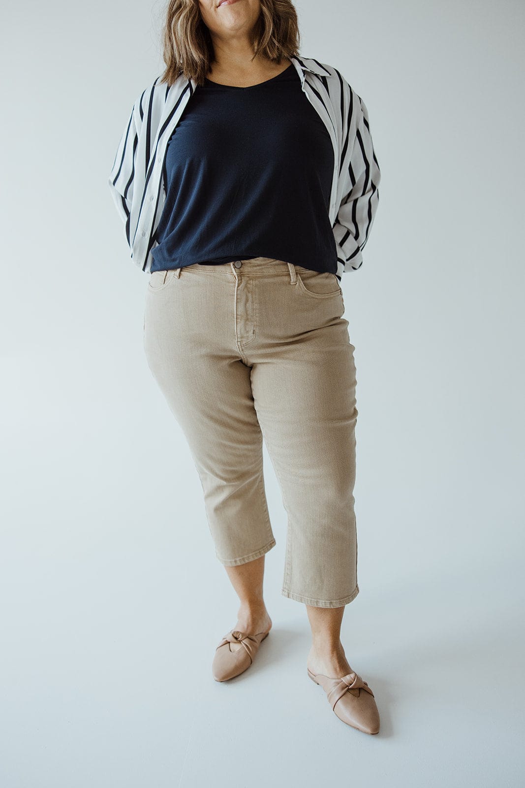 A person wearing a black top, black and white striped jacket, Judy Blue JUDY BLUE NON-DISTRESSED CAPRI IN WASHED KHAKI, and beige shoes stands against a plain background, showcasing an effortlessly chic summer style.