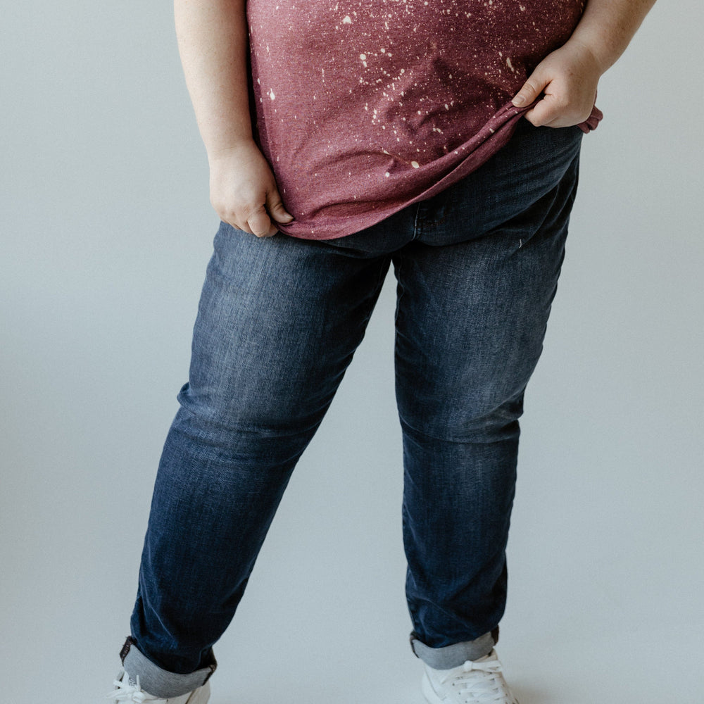 A person dressed in a maroon shirt, cuffed slim fit jeans, and white sneakers is standing against a plain background.