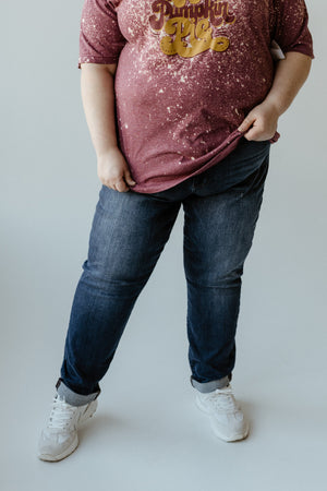 A person dressed in a maroon shirt, cuffed slim fit jeans, and white sneakers is standing against a plain background.