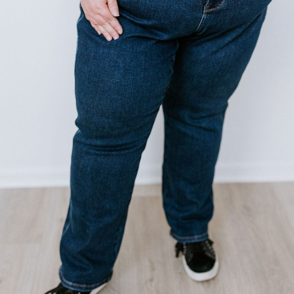 A person wearing a brown shirt, Judy Blue Straight Leg Tummy Control Dark Wash Jeans, and black sneakers is standing on a light-colored wooden floor.