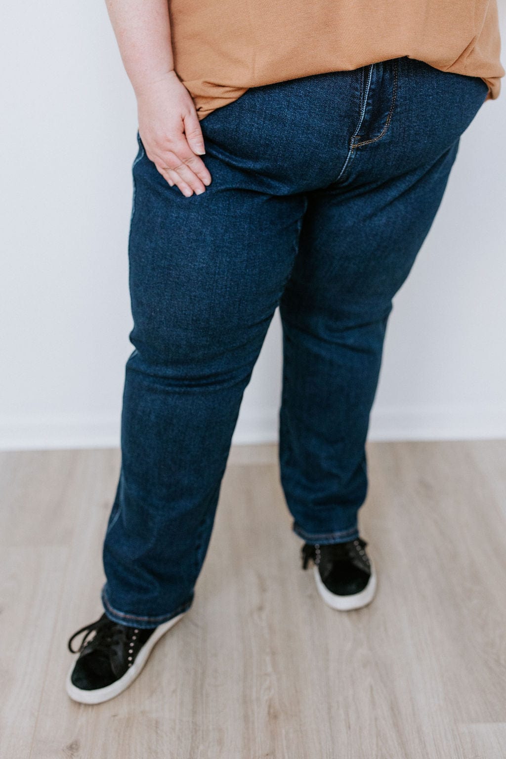 A person wearing a brown shirt, Judy Blue Straight Leg Tummy Control Dark Wash Jeans, and black sneakers is standing on a light-colored wooden floor.