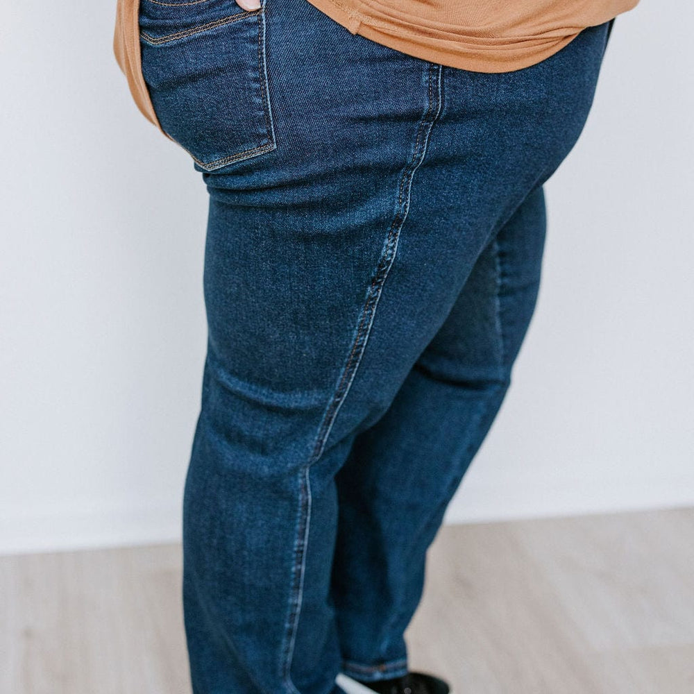 Side view of a person wearing a brown shirt, JUDY BLUE STRAIGHT LEG TUMMY CONTROL DARK WASH JEANS by Judy Blue, and black sneakers, with one hand placed in the back pocket.