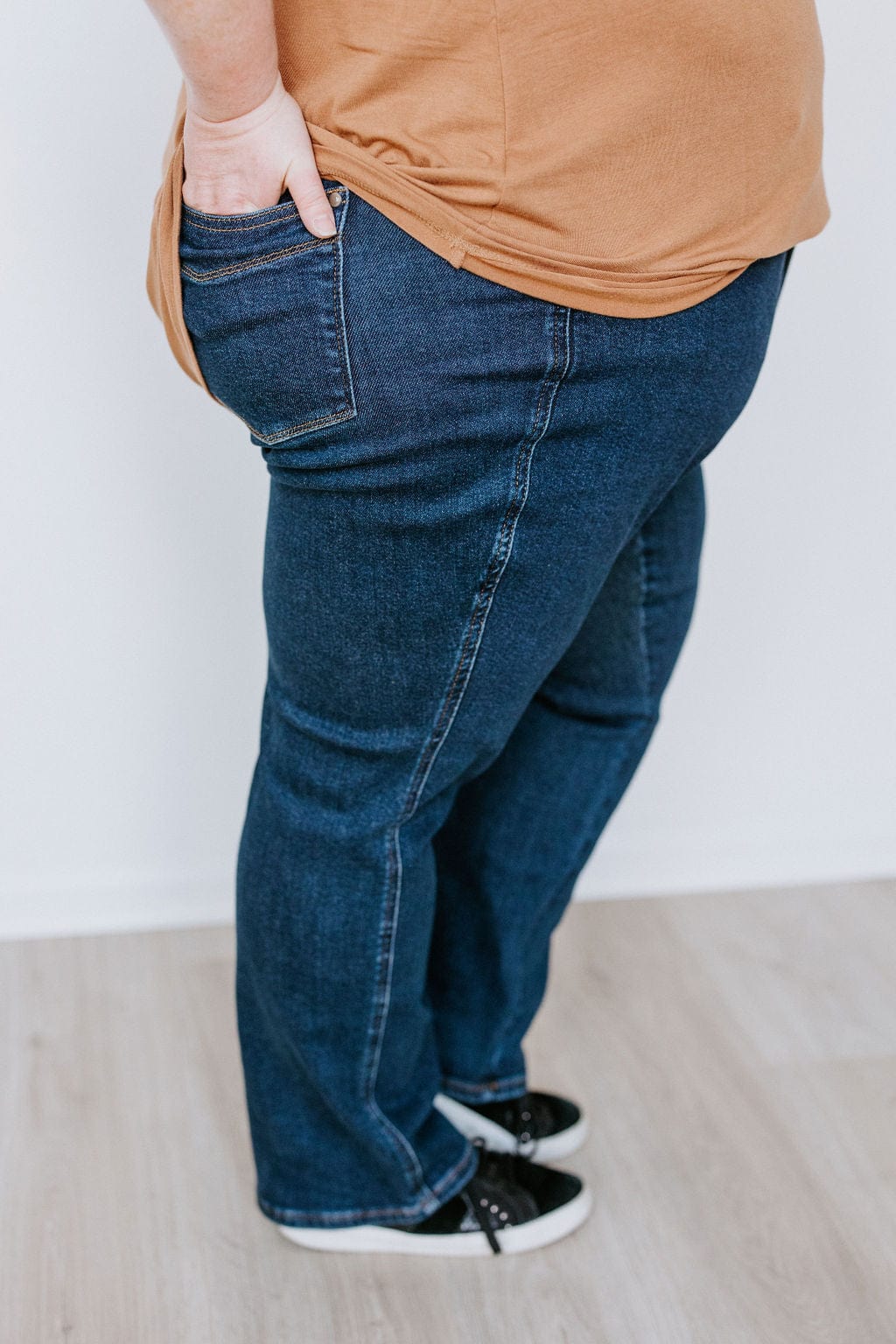 Side view of a person wearing a brown shirt, JUDY BLUE STRAIGHT LEG TUMMY CONTROL DARK WASH JEANS by Judy Blue, and black sneakers, with one hand placed in the back pocket.