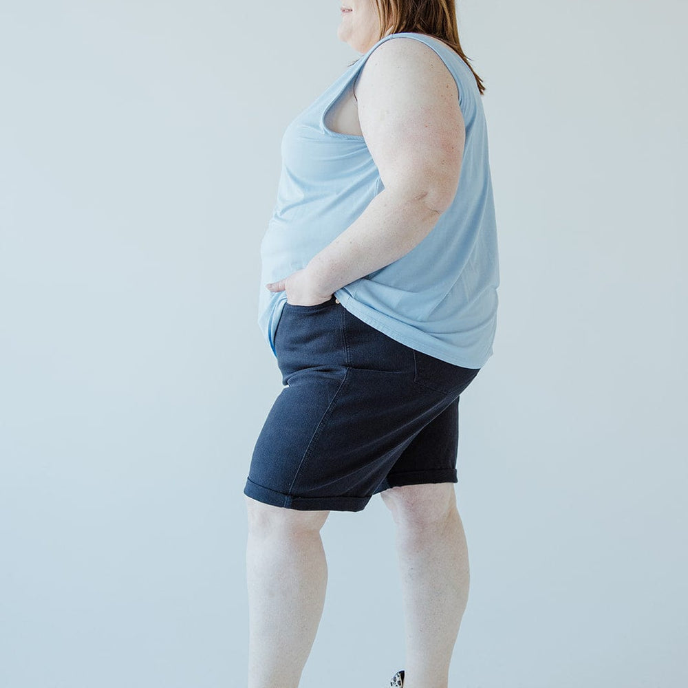 A person stands sideways with hands in pockets, wearing a light blue sleeveless top, JUDY BLUE TUMMY CONTROL BERMUDA SHORTS IN NAVY by Judy Blue, and light-colored sneakers against a plain white background.