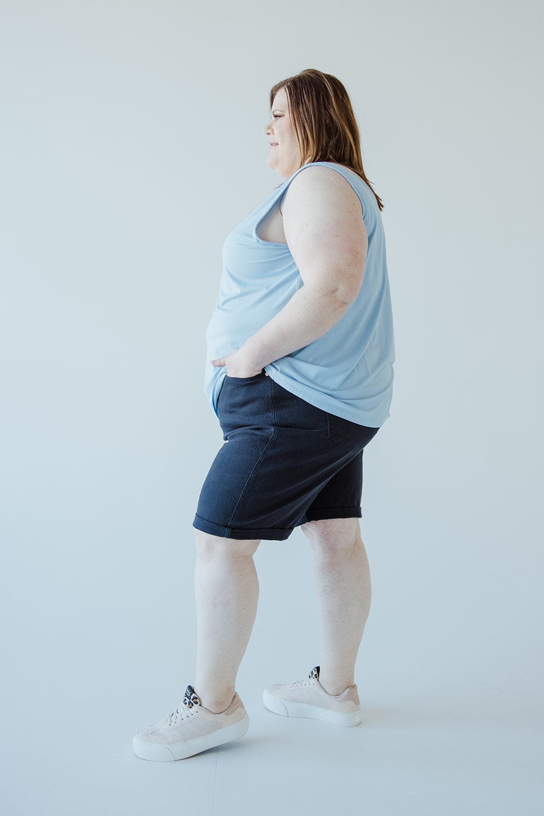 A person stands sideways with hands in pockets, wearing a light blue sleeveless top, JUDY BLUE TUMMY CONTROL BERMUDA SHORTS IN NAVY by Judy Blue, and light-colored sneakers against a plain white background.