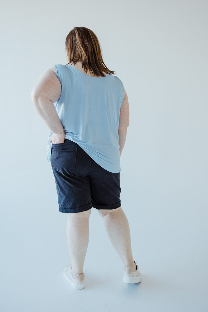 Woman wearing a light blue shirt, JUDY BLUE TUMMY CONTROL BERMUDA SHORTS IN NAVY from Judy Blue, and white shoes, standing with her back to the camera against a plain, light grey background.