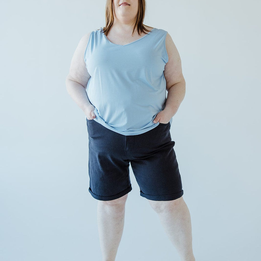 A woman stands against a plain backdrop, wearing a light blue sleeveless top, JUDY BLUE TUMMY CONTROL BERMUDA SHORTS IN NAVY by Judy Blue, and pink sneakers. She is looking slightly upwards with her hands in her pockets.