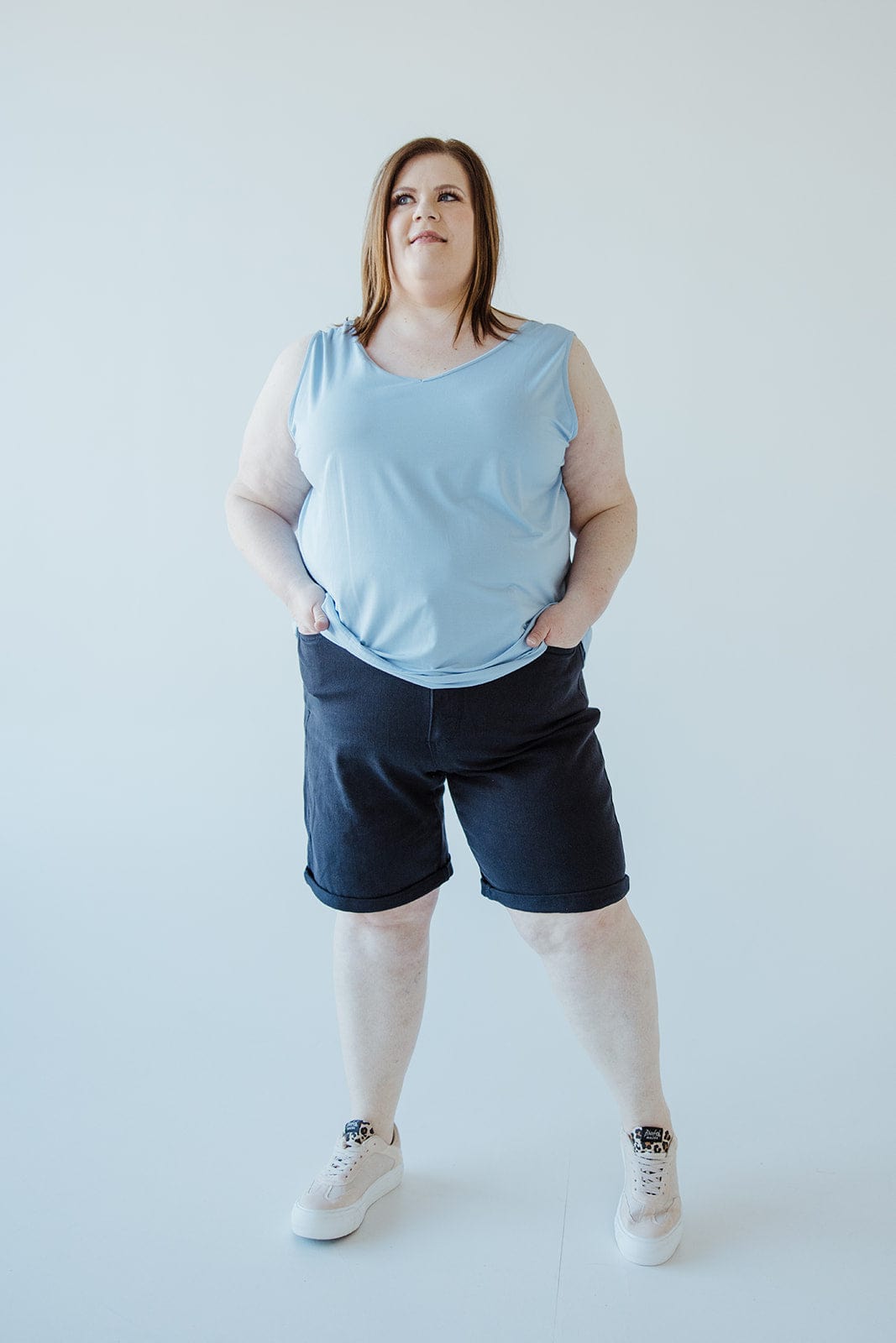 A woman stands against a plain backdrop, wearing a light blue sleeveless top, JUDY BLUE TUMMY CONTROL BERMUDA SHORTS IN NAVY by Judy Blue, and pink sneakers. She is looking slightly upwards with her hands in her pockets.