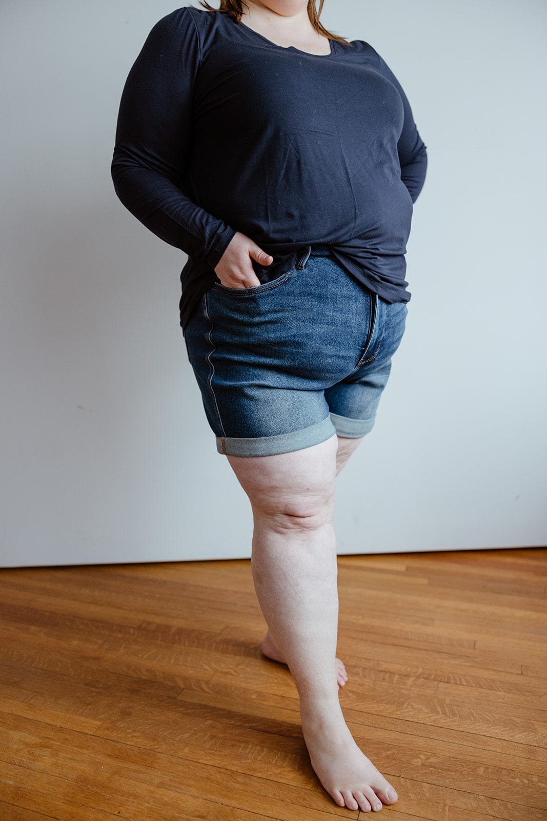 Person in a black long-sleeve shirt standing barefoot on a wooden floor, sporting Judy Blue JUDY BLUE TUMMY CONTROL CUFFED SHORTS IN VINTAGE WASH with hands in pockets.