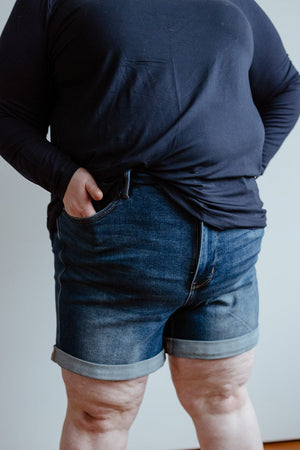 Person wearing a dark long-sleeve shirt and Judy Blue JUDY BLUE TUMMY CONTROL CUFFED SHORTS IN VINTAGE WASH with a high-rise adjustable inseam, hands partially in the shorts' pockets.