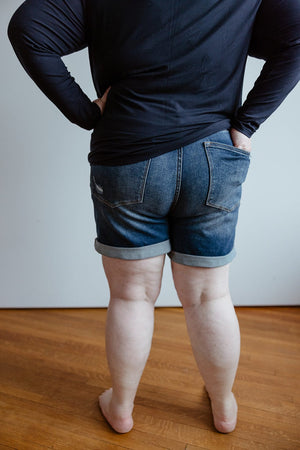 Person standing with their back to the camera, wearing a black long-sleeve shirt and Judy Blue Tummy Control Cuffed Shorts in Vintage Wash by Judy Blue, against a plain background.